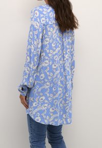 Blouse florale bleu clair à manches longues, avec des motifs floraux blancs et une coupe décontractée, associée à un jean en denim bleu.