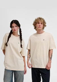 Beige oversized t-shirts lavet af blødt bomuld, med et lille grønt logo på brystet. Den mandlige model bærer mørke bukser; den kvindelige model bærer jeans.