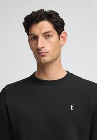 Svart sweatshirt i slätt tyg, med rund halsringning och en liten vit logotyp av en figur med en golfklubba på vänster bröst.