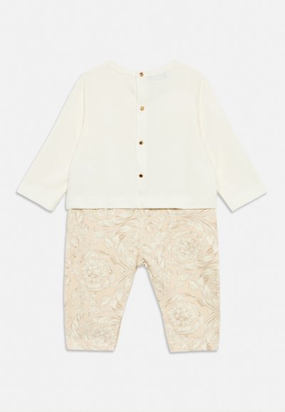 Krém színű hosszú ujjú babaruházat arany gombokkal a háton és bézs virágmintás nadrággal rögzítve.