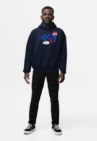 Uomo che indossa una felpa navy blue con il logo della Pepsi, pantaloni neri con motivo a griglia bianca e sneakers in bianco e nero, in piedi su uno sfondo bianco.