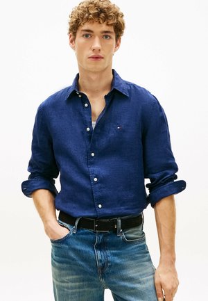 Ung mand med krøllet hår iført en mørkeblå skjorte med knapper og op-rullede ærmer samt lyseblå jeans, stående med den ene hånd i lommen.