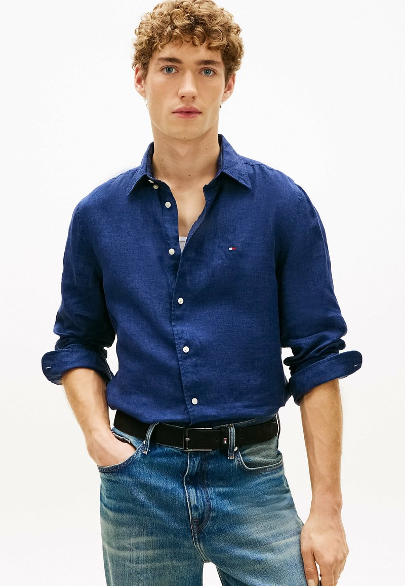 Jeune homme aux cheveux bouclés portant une chemise bleu foncé à boutons avec les manches retroussées et un jean bleu clair, debout avec une main dans la poche.