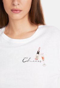Weißes T-Shirt mit bestickter Champagnerflasche mit einer schwarzen Schleife und einem Glas Champagner, begleitet von dem Text "Prost" in geschwungener Schrift.