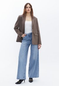 Bruine blazer, witte top en lichtblauwe wijde high-waisted spijkerbroek. Zwarte puntige hakken met kettingdetail. Eenvoudige achtergrond.