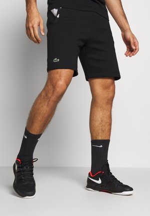 Homme portant un short noir Lacoste, des chaussettes Nike noires et des chaussures de sport Nike noires avec des touches rouges, debout sur un sol clair.