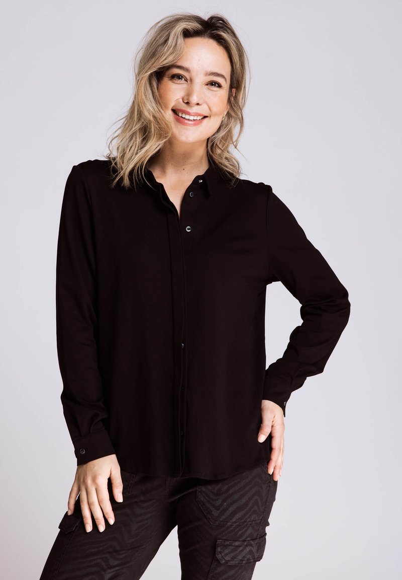 Schwarzes langärmliges Hemd mit Knöpfen und Kragen, mit glatter Textur und subtiler Schimmer, kombiniert mit dunklen, gemusterten Cargohosen.