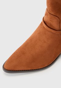 Bottine en suède marron avec un bout pointu, présentant une semelle foncée contrastante et un détail de sangle enroulée au niveau de la cheville.