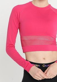Rosa langärmliger Cropped-Top mit Mesh-Mittelteil, getragen von einer Person mit Champion-Logo am Bund der schwarzen Leggings.