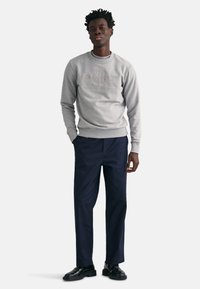 Grijze sweatshirt met reliëflogo, gecombineerd met marineblauwe broek. Model draagt zwarte schoenen. Zacht materiaal, ronde hals, comfortabele pasvorm, minimalistisch ontwerp.