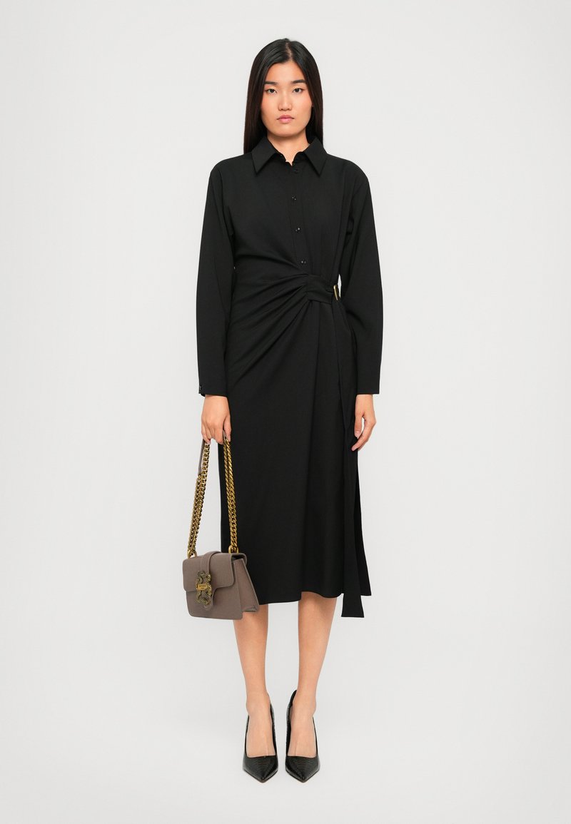 Vrouw die staat in een zwart midi-jurk met lange mouwen en een zij-knoopdetail, zwarte hakken, en een taupe handtas met gouden kettinghengsels vasthoudt.
