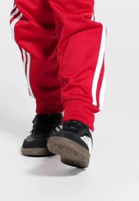 Čierne športové topánky s bielymi pruhmi, kombinované s červenými teplákmi so svetlými bočnými pruhmi a rebrovanými manžetami.