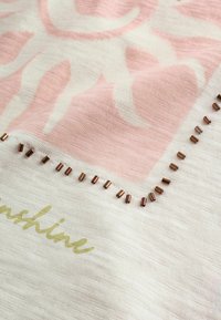 Nahaufnahme von weißem und rosa Stoff mit metallischen Perlen entlang einer Naht und teilweise sichtbaren, goldfarbenen Schreibschriftbuchstaben.