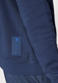 Sudadera de Nike en azul marino con puños y dobladillo acanalados, que presenta una etiqueta azul con texto y logo en el lateral. Textura suave, diseño casual.