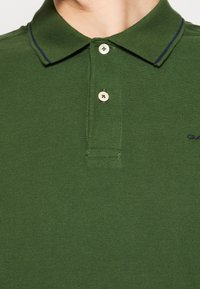 Homme portant un polo vert foncé avec deux boutons blancs et des rayures bleu marine sur le col, gros plan sur la poitrine et le cou.