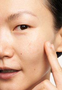 Primo piano di un volto con pelle liscia e chiara, decorata da qualche lentiggine. Un dito sfiora delicatamente un'area lucida e idratata sulla guancia.