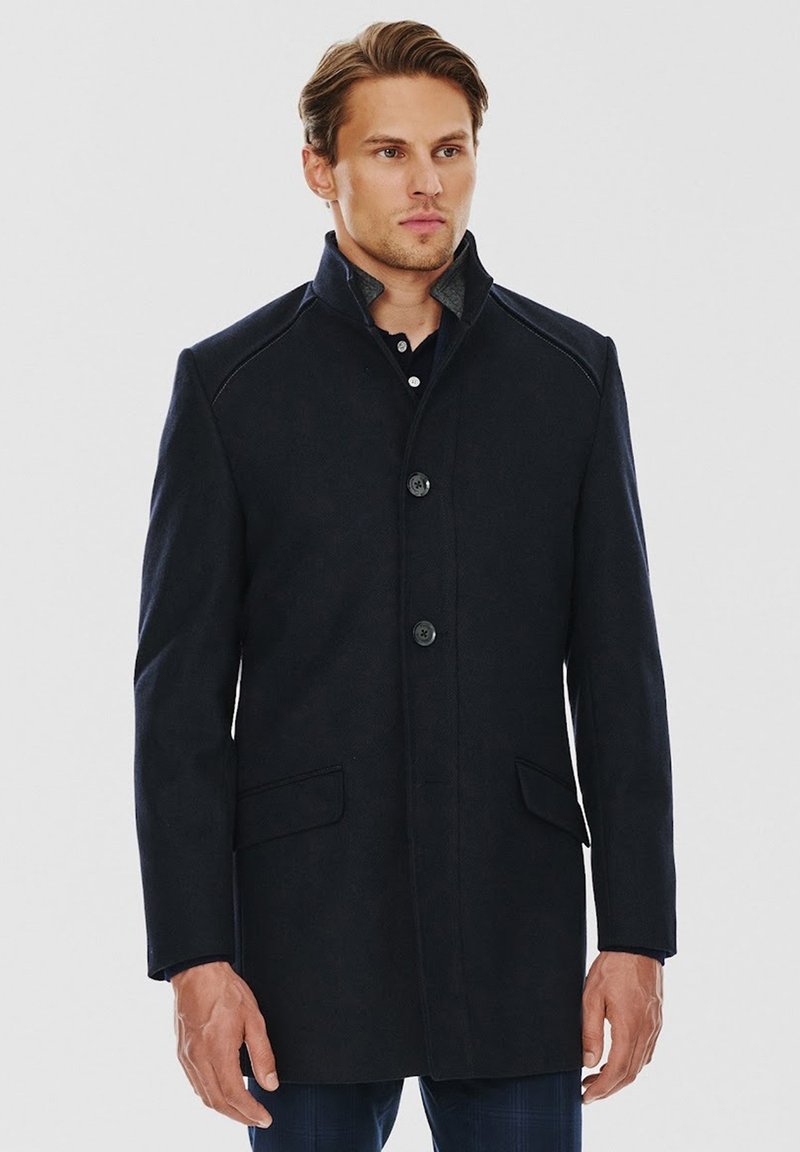 Granatowy wełniany płaszcz o prostym kroju, z stójką, dwiema przednimi kieszeniami i zapięciem na guziki. Posiada subtelne przeszycia na ramionach oraz fakturowany materiał.