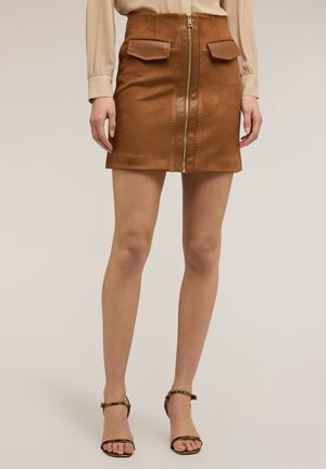 Kobieta w brązowej zamszowej mini spódnicy z przednim zamkiem błyskawicznym i kieszeniami z klapami, beżowej bluzce z długim rękawem oraz brązowych szpilkach z paskiem wokół kostki.