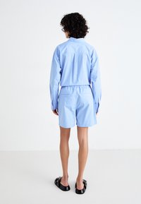 Chemise bleu clair à manches longues avec des poignets à boutons et une taille élastique. Associée avec un short assorti et des sandales noires à glisser.