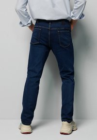 Donkerblauwe denim jeans met een klassieke pasvorm, subtiele stiksels en twee achterzakken, gecombineerd met lichtgekleurde sportschoenen.