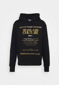 Černá mikina s kapucí se zlatým nápisem "Versace Jeans Couture" a dekorativními grafikami řetězu na hrudi, obsahující přední kapsu a stahovací kapuci.