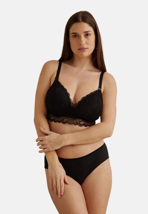 Femme aux longs cheveux bruns portant un soutien-gorge en dentelle noire avec des bords festonnés et une culotte noire assortie sur un fond blanc.