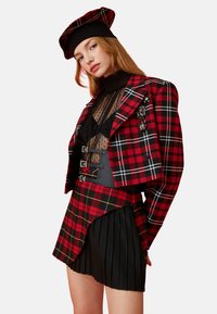 Rood en zwart geruite cropped jas met zilveren hardware, gelaagd over een doorschijnende zwarte top, gecombineerd met een geplooide zwarte rok en bijpassende hoed.