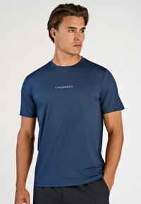 Mand iført en marineblå kortærmet T-shirt med "LINDBERGH" trykt på brystet, stående med den ene hånd i lommen og kiggende til siden.