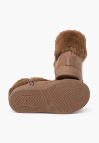 Calzado en tono beige con puño de pelo sintético, parte superior de ante y suela de goma que presenta un patrón de tracción en zigzag. Cierre lateral con cremallera para facilitar su uso.