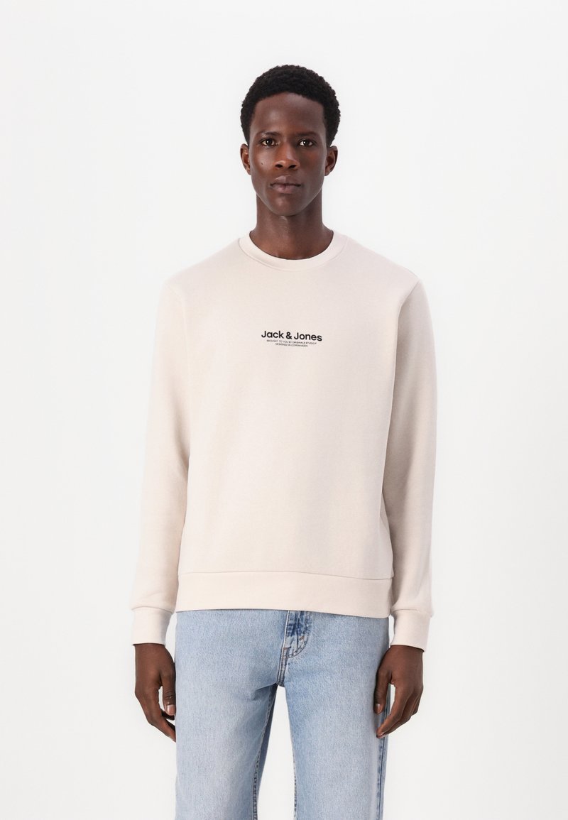 Man in een beige Jack & Jones sweatshirt en lichtblauwe jeans, staand tegen een effen witte achtergrond, recht naar voren kijkend.