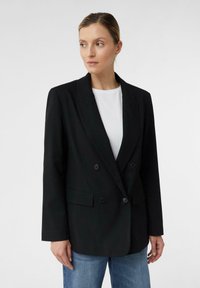 Donna che indossa un blazer nero doppiopetto sopra una camicia bianca e dei jeans blu, che guarda leggermente di lato su uno sfondo neutro.