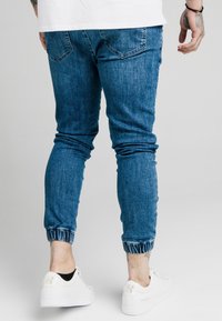 Jeans de mezclilla azul ajustados con piernas entalladas, puños elásticos y bolsillos traseros. Llevados con zapatillas blancas con detalles dorados.