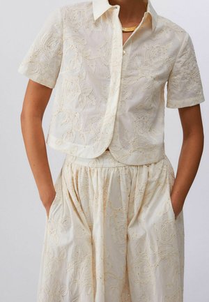 Blouse beige à manches courtes boutonnée et jupe assortie avec broderie florale, mains du modèle dans les poches de la jupe.