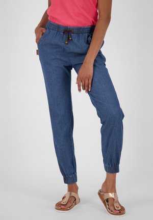 Kvinde iført blå joggebukser med elastisk talje og rosaguldsandaler, står med den ene hånd i lommen, og en pink skjorte er synlig.