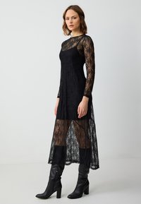 Zwarte kanten jurk met lange mouwen, doorschijnende overlay en midi-lengte stijl. Gecombineerd met kniehoogte zwarte laarzen. Elegante bloemenpatroon detail.