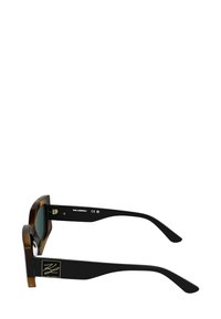 Okulary przeciwsłoneczne w kolorze muszli żółwia z masywną oprawką, ciemnymi szkłami i czarnymi zausznikami z złotym logo po bokach. Gładka tekstura, wyrazisty design.