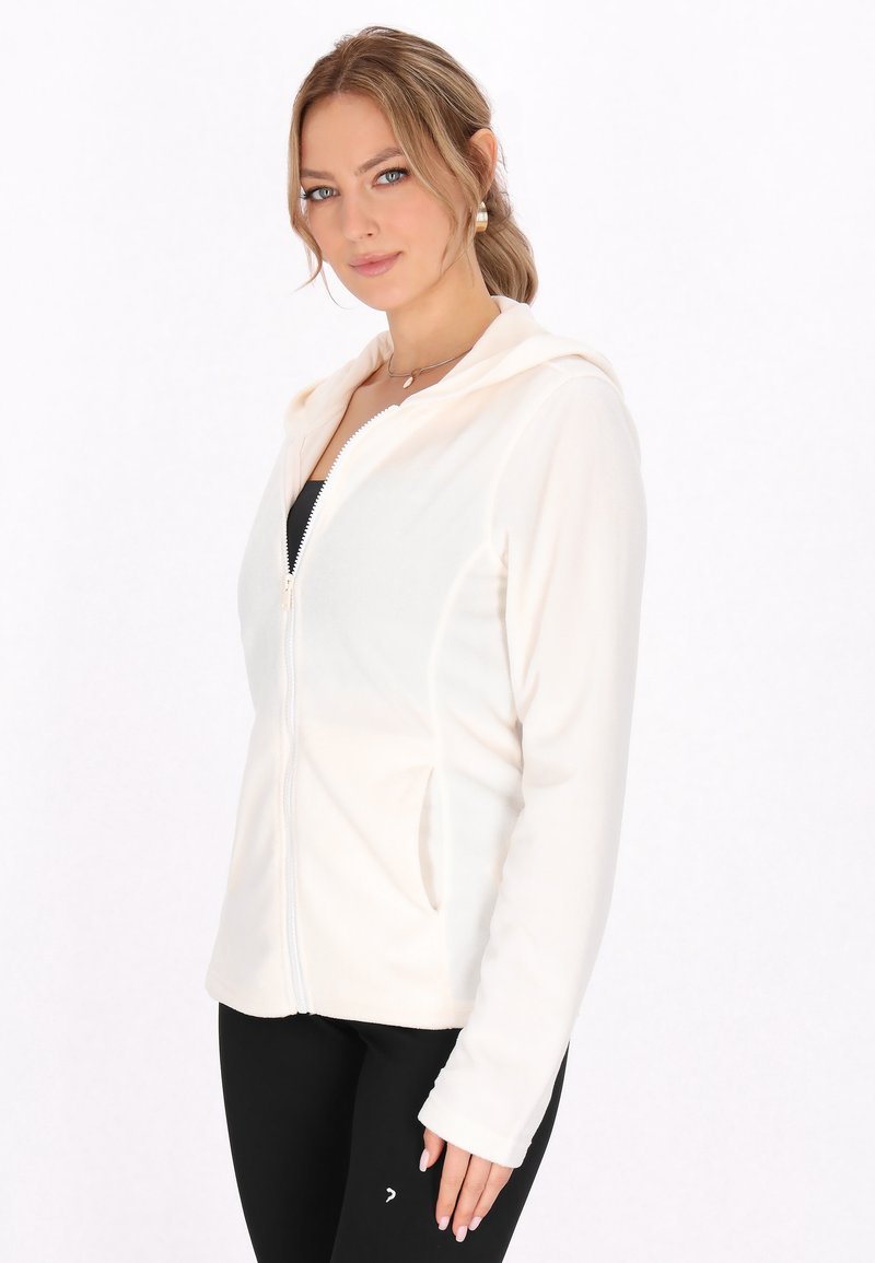 Helle Creme Zip-Hoodie aus weichem Stoff mit Kapuze, seitlichen Taschen und langen Ärmeln, kombiniert mit schwarzen Leggings. Einfaches Design.
