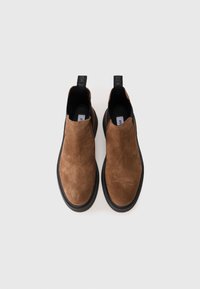 Zapatos slip-on de ante marrón con suela de goma negra, con una textura suave y una lengüeta para facilitar su uso. Diseño minimalista.