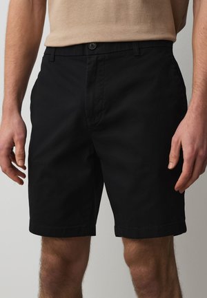 Mand iført sorte knælange shorts og en beige t-shirt med hænderne afslappet langs siden mod en ensfarvet baggrund.