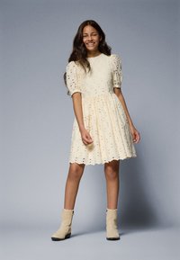 Robe couleur crème avec des manches bouffantes, des détails en broderie anglaise et une jupe évasée. Associée à des bottines beige clair.