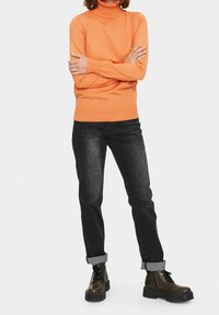 Orange turtlenecktröja med långa ärmar, i kombination med svarta jeans och mörka kängor. Texturen är slät, med uppvikta ärmslut på byxorna.