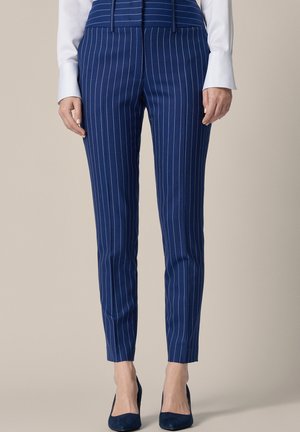 Kobieta ubrana w niebieskie, dopasowane spodnie w prążki z wysokim stanem oraz ciemnoniebieskie szpilki, stojąca na tle jednolitego beżowego tła.