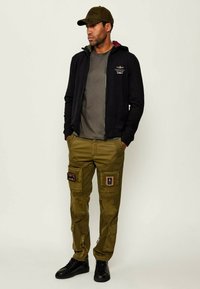 Felpa nera con zip, pantaloni cargo verde oliva con toppe, t-shirt grigia a girocollo e scarpe da ginnastica nere. Il cappellino abbina la felpa.