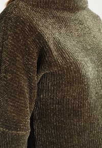 Pull en maille texturée et à côtes de couleur vert olive foncé. Il présente un col haut et une coupe ample, avec une douceur et une profondeur visibles dans le tissu.