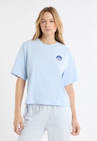 Jasnoniebieski bawełniany t-shirt z krótkimi rękawami, okrągłym dekoltem i haftowaną okrągłą naszywką z wzorem. Krótki fason powyżej wzorzystych spodni.