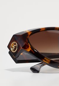 Occhiali da sole tartarugati con lenti angolari marrone scuro, accento con logo dorato sul lato e asta nera elegante.
