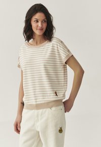 Haut beige rayé à manches courtes, avec un ourlet côtelé et des détails en boutons. Présente des accents brodés près du bas. Associé à un pantalon blanc.