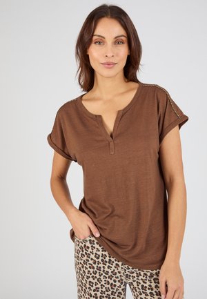 Frau trägt ein braunes Kurzarmshirt mit abgestepptem Schulterdetail und Leopardenhose, steht mit einer Hand in der Hosentasche vor weißem Hintergrund.