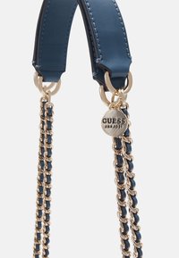 Bandoulière de sac en cuir bleu avec une chaîne dorée et un charme rond en or gravé du nom de la marque Guess et de l’année de création.
