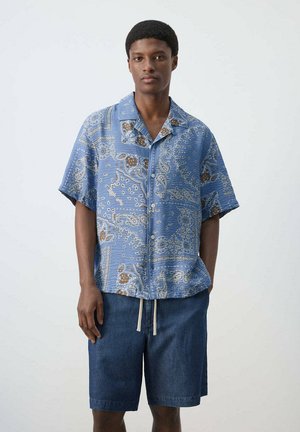 Joven con camisa azul de manga corta con estampado paisley y pantalones cortos vaqueros, de pie contra un fondo claro y liso.